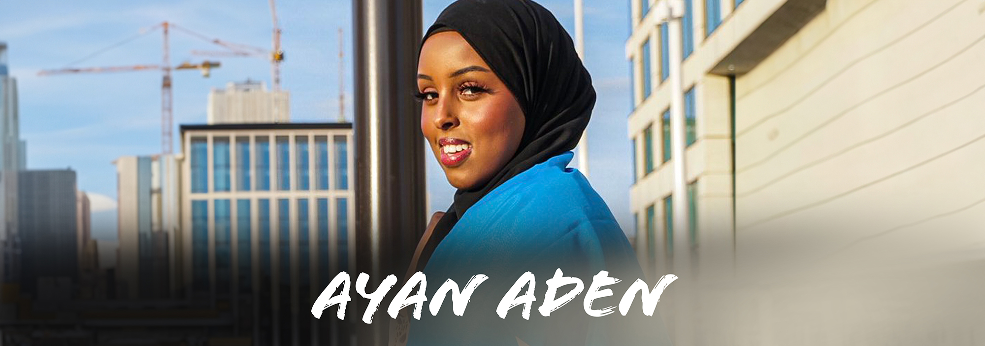 A young female of dual Somali / English identity, wearing a head scarf and she has the Somali flag draped around her shoulders. She is standing in front of a modern city background with a bright blue sky. She is smiling and facing the camera.