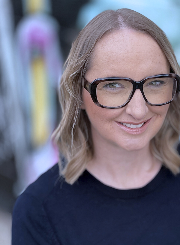 Female in her early 40’s with fair skin, shoulder length wavy hair, wearing a navy jumper and large dark glasses. She is smiling at the camera standing in front of a colourful concrete wall outside.