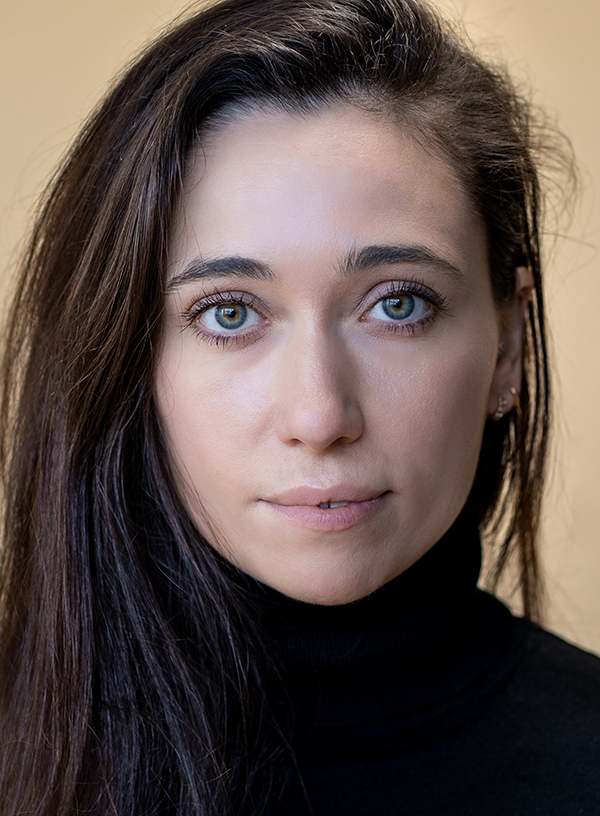 Image description: headshot of young fair-skinned adult female looking straight at the camera in a studio setting with long dark brown straight hair. She has a neutral expression and is wearing a high-necked black jumper.