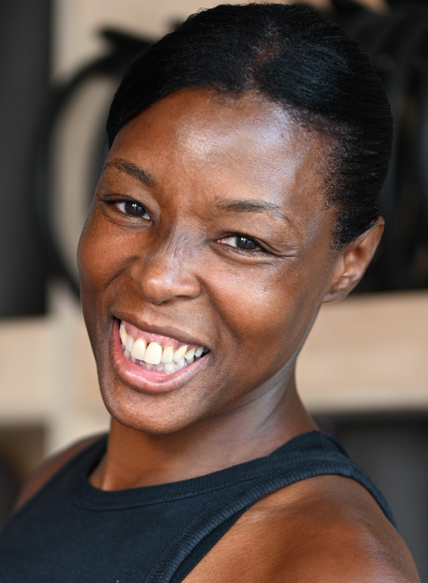 headshot of a dark-skinned female adult with a broad smile, black Afro hair tied back, wearing a black round-necked vest top in a studio setting.