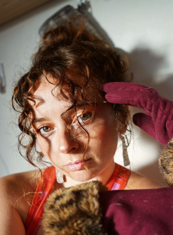 A headshot of a white woman in her 20s/30s, with brown curly hair tied up, loose curls falling in front of her face. She is looking at the camera, has brown eyes and is wearing a pink/orange vest top and maroon gloves with fur edges.