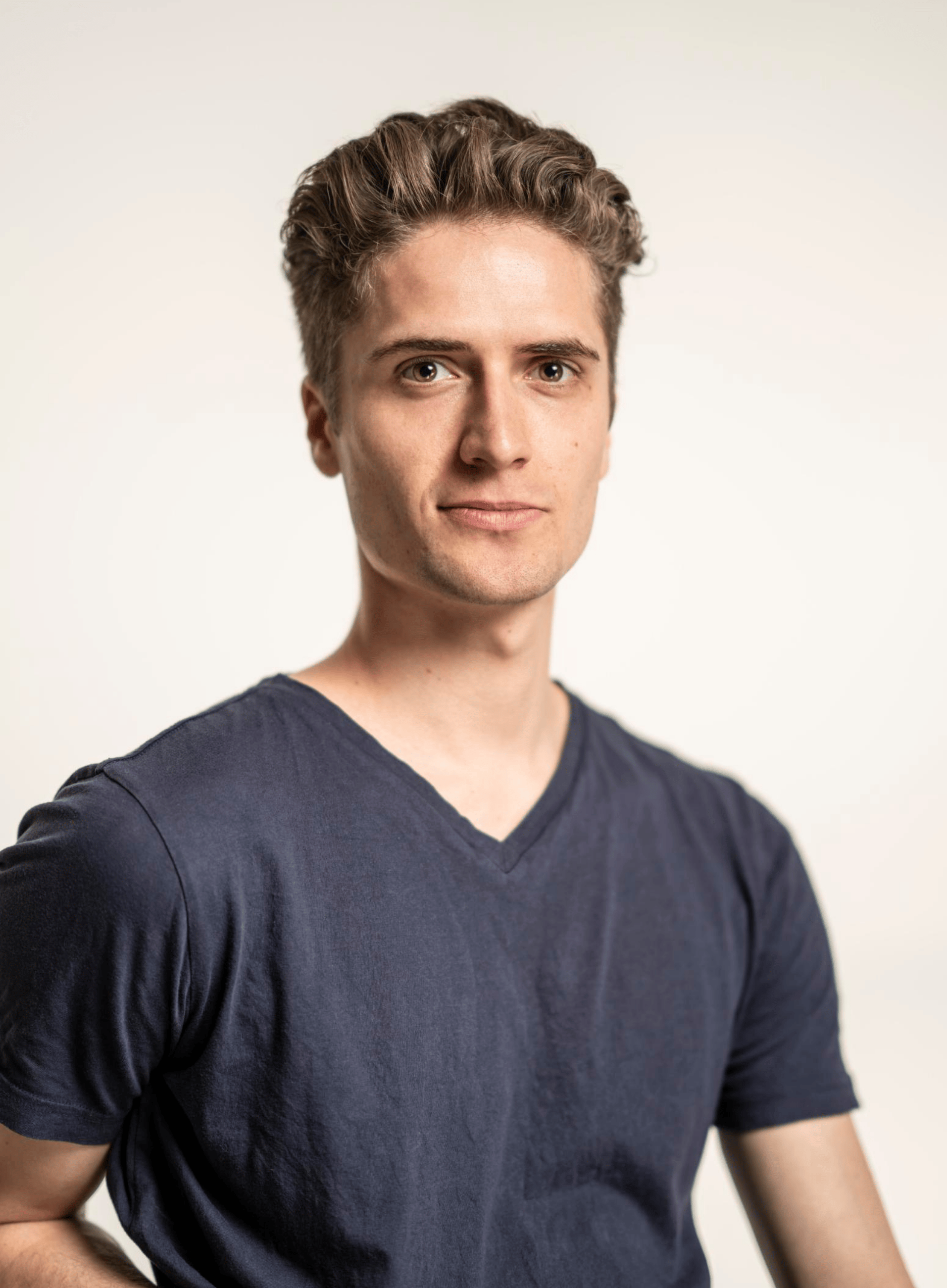 A head and shoulders photo of August Generalli, a fair-skinned man in his 30s with brown hair, wearing a blue v-necked t-shirt