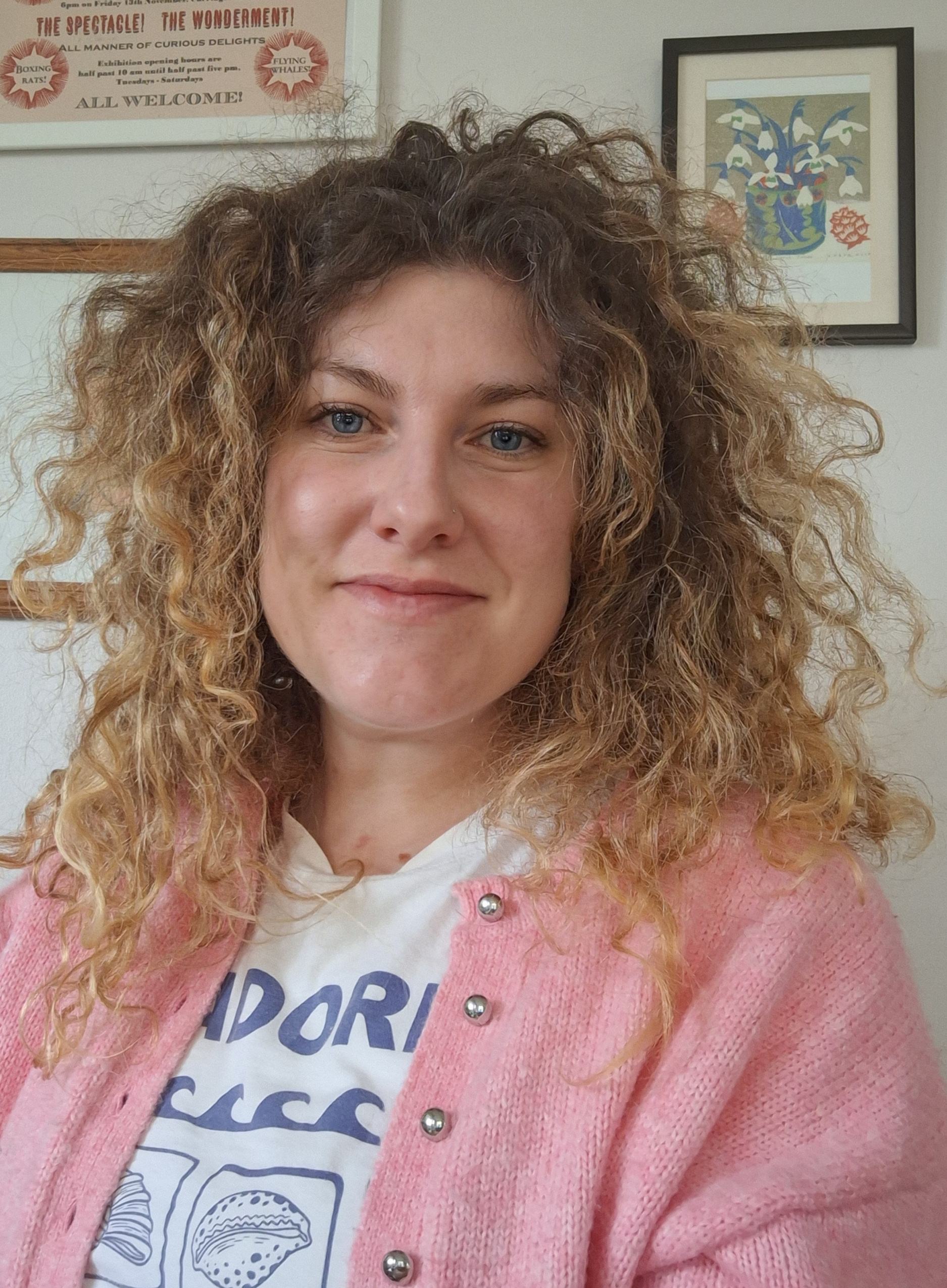 Headshot of Jen Warner, a fair skinned woman in her 30s with long, curly blonde/brown hair, wearing a pink cardigan over a white t-shirt and smiling towards the camera.