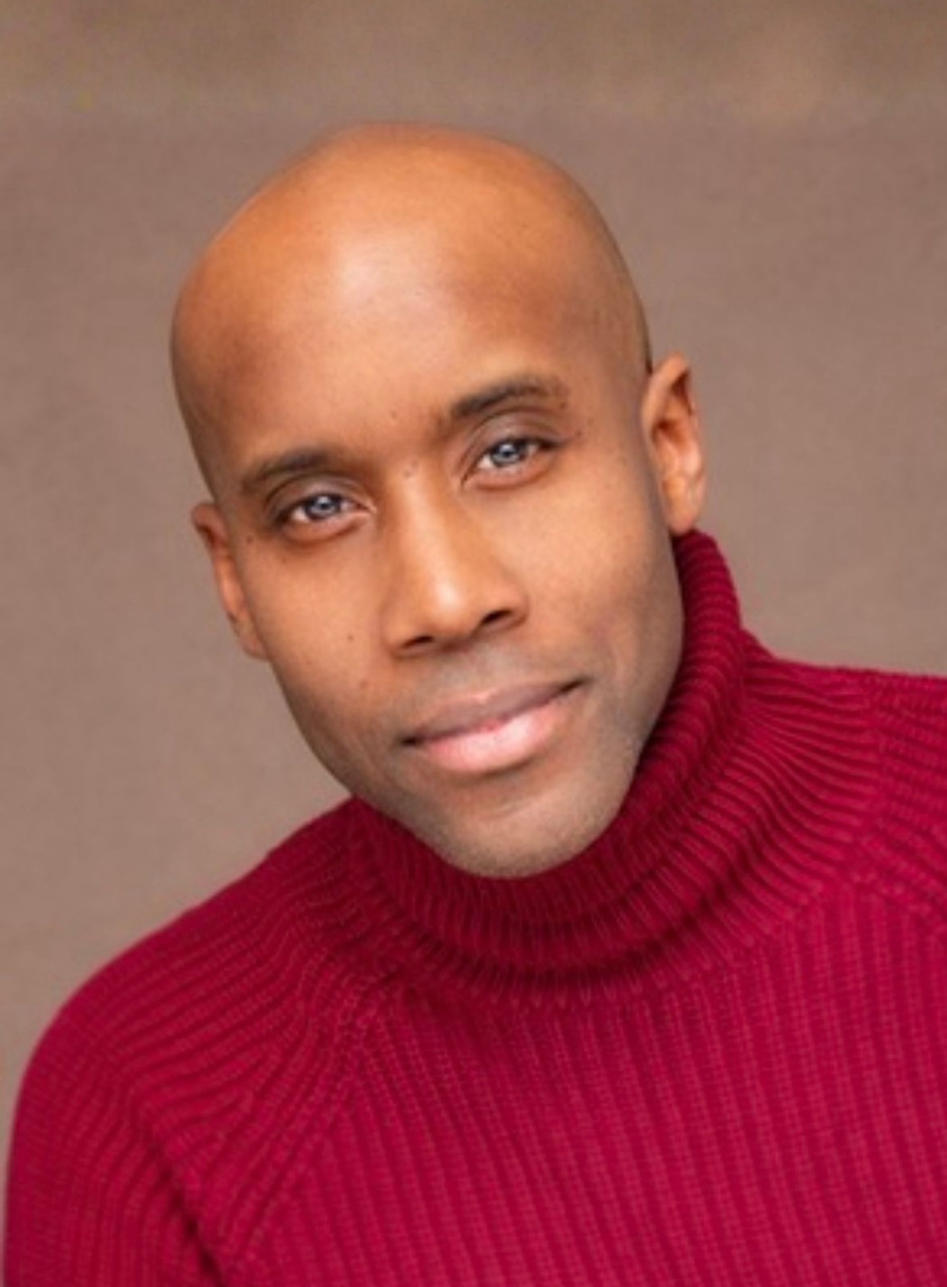 Headshot of a man with dark brown skin and a shaved head, tilting his head slightly with a gentle, closed-mouth smile. He has light-coloured eyes and wears a deep red ribbed roll-neck jumper. The background is a soft, warm brown.