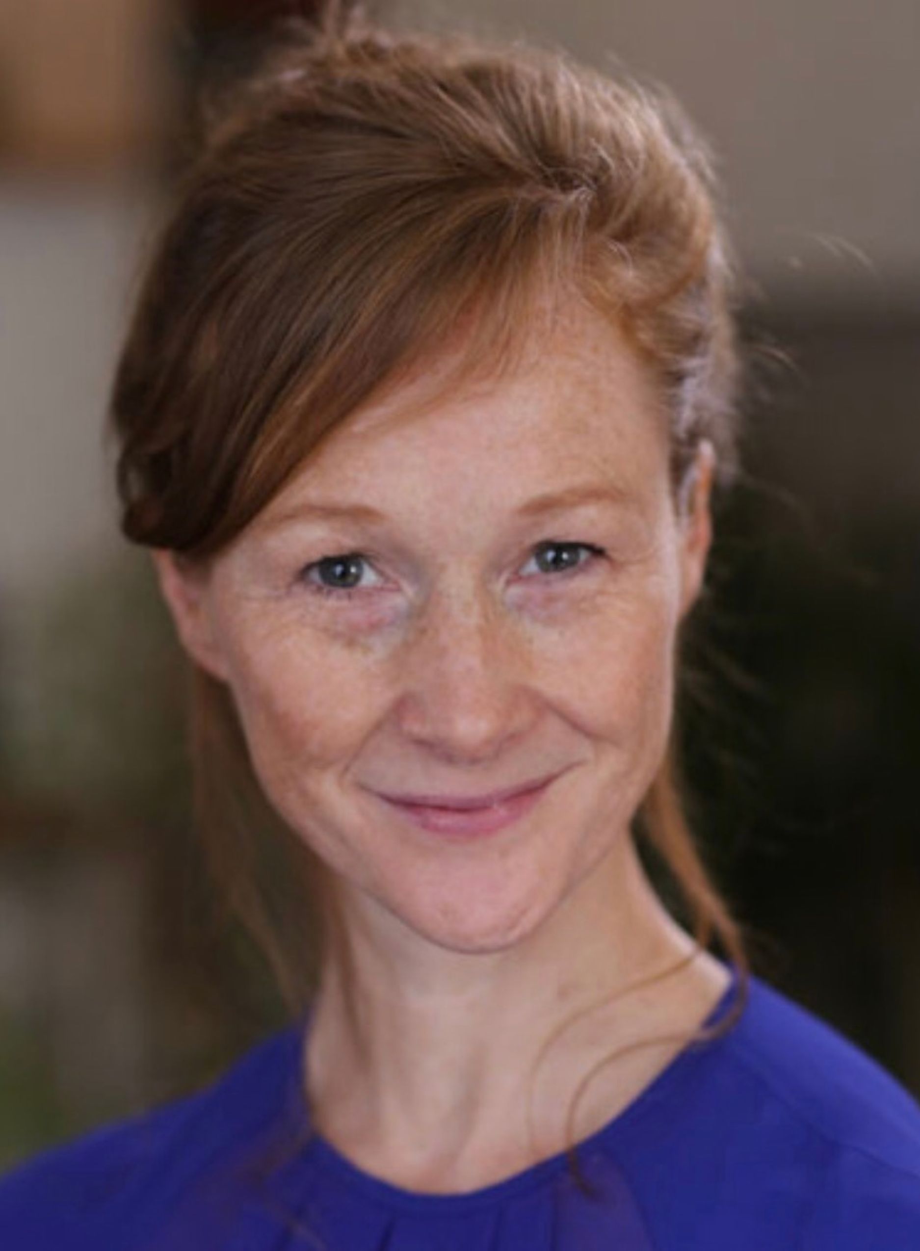 Headshot of a woman with fair, freckled skin and auburn hair swept up and to one side. She has blue-grey eyes and smiles warmly at the camera. She wears a bright blue top against a softly blurred background.